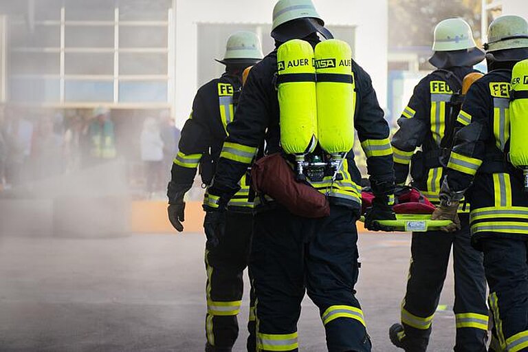 Vier Feuerwehrleute tragen in voller Schutzkleidung ihre Ausrüstung in Richtung eines stark rauchenden Gebäudes.