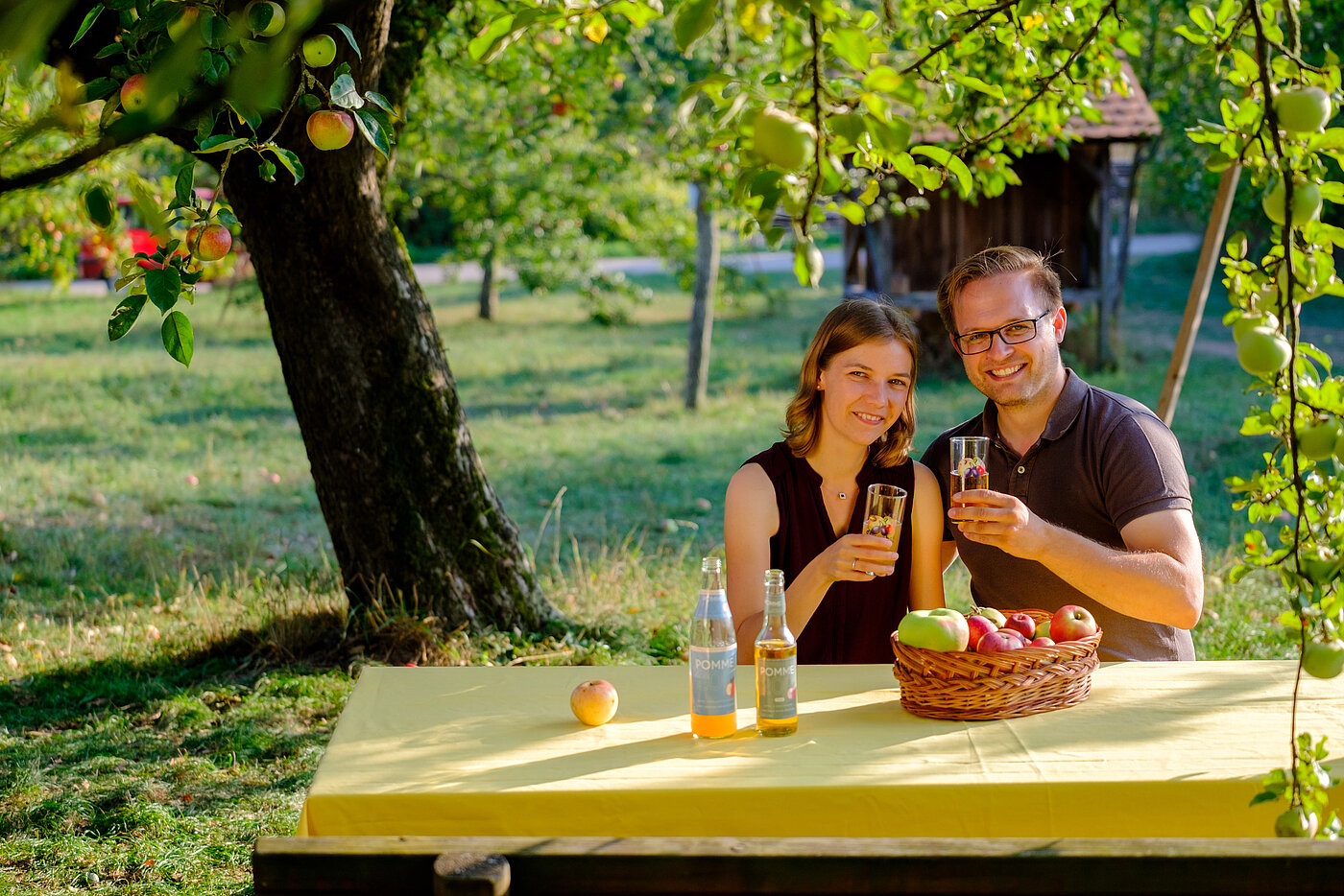 Ein junges Parr sitzt an einem Tisch unter einem Apfelbaum. Sie stoßen mit einem Glas Apfelsaft an und lächeln in die Kamera.