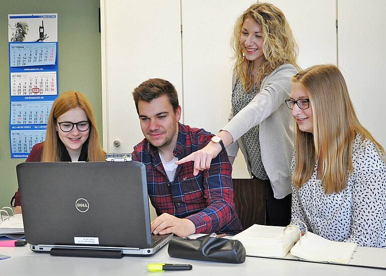 Vier Auszubildende vor einem Laptop. Drei sitzen, eine Auszubildende steht und deutet auf den Laptop-Bildschirm.
