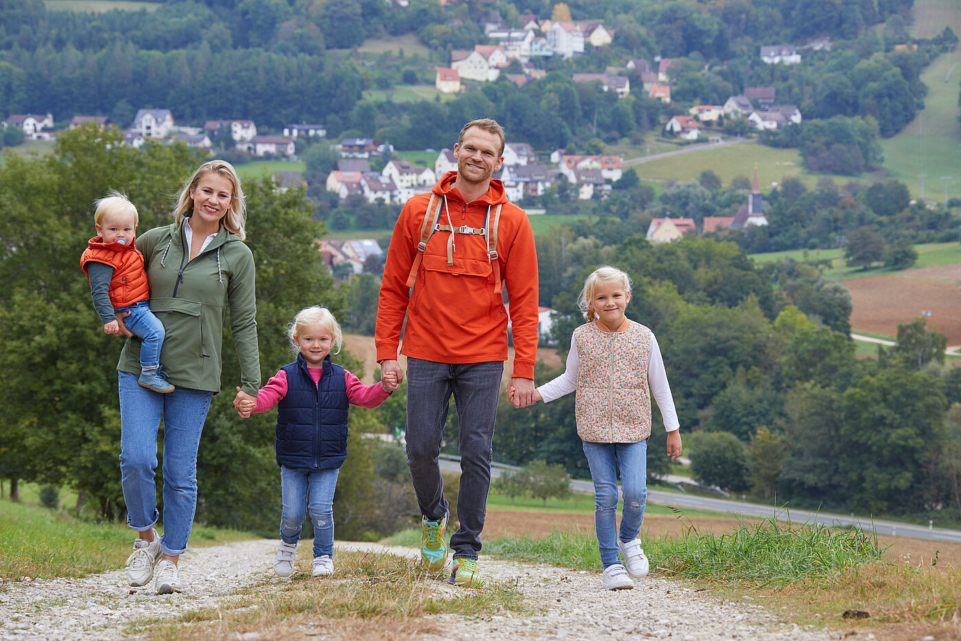 Eine fünfköpfige Familie in Outdoor-Bekleidung weit entfernt Wälder und Dörfer. spaziert Hand in Hand über einen Hügel. Im Hintergrund sieht man 