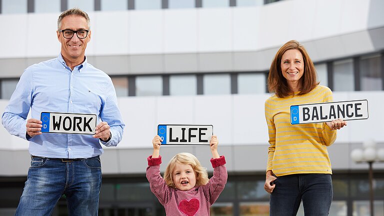 Ein Mann, ein kleines Mädchen und eine Frau stehen vor dem Landratsamt, lachen in die Kamera und halten jeweils ein Schild hoch. Darauf stehen die Worte "Work", "Life" und "Balance".
