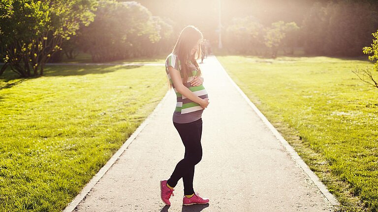 Eine junge Frau steht auf einem von der Sonne beschienenen Weg im Grünen. Sie hält mit beiden Händen ihren Schwangerschaftsbauch.