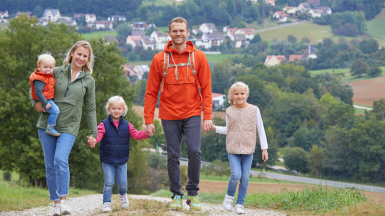 Eine fünfköpfige Familie in Outdoor-Bekleidung weit entfernt Wälder und Dörfer. spaziert Hand in Hand über einen Hügel. Im Hintergrund sieht man 