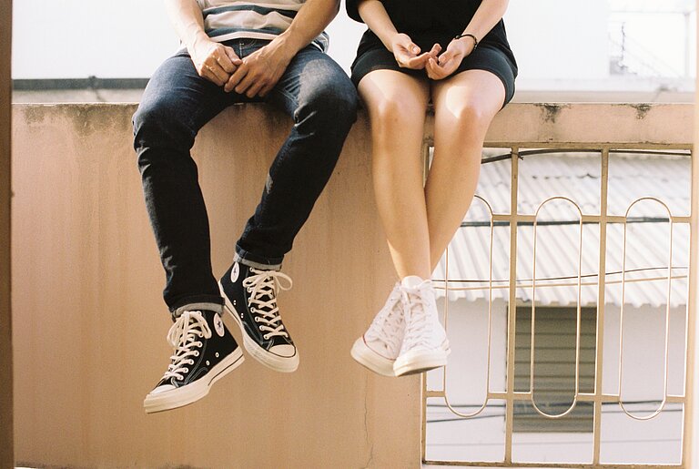 Eine Mauer, auf der ein junger Mann und eine junge Frau eng nebeneinander sitzen und ihre Beine baumeln lassen. Beide sind nur vom Bauch abwärts zu sehen. 