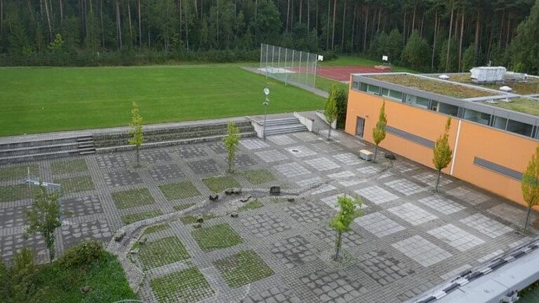 Außenansicht der Realschule Röthenbach: Vor einem orangenen Gebäude sind auf einem grauen Platz einige Bäume gepflanzt worden, im Hintergund sind ein Fußballplatz und ein Wald zu sehen.