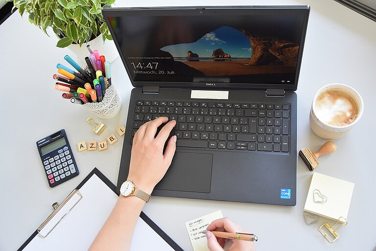 Laptop mit Startbildschirm umgeben von Schreibtischutensilien. Eine Hand tippt gerade auf der Tastatur.