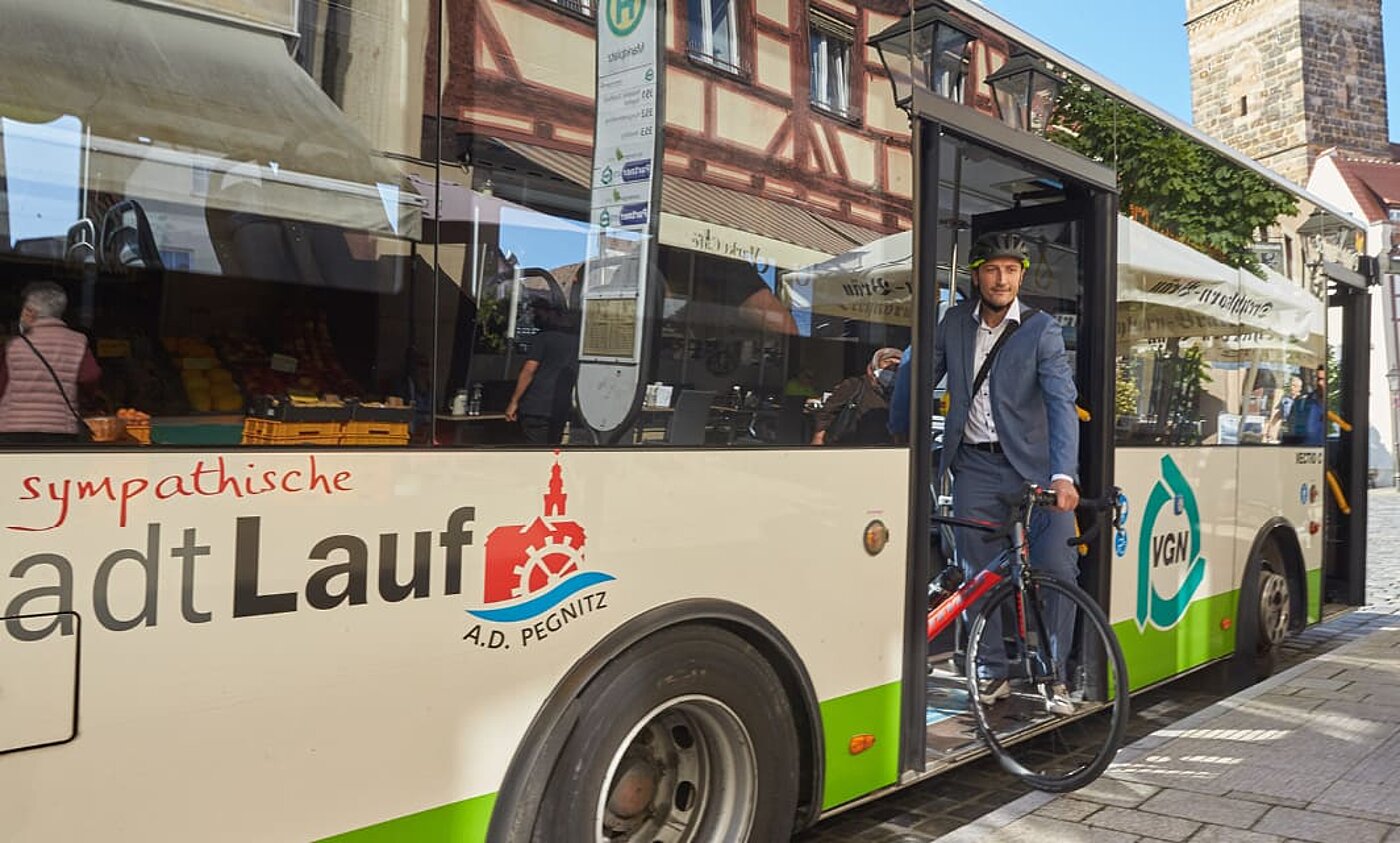 Ein Mann im Anzug hebt sein Rennrad aus einem Linienbus. Er trägt einen Fahrradhelm. Auf dem weiß-grünen Bus steht "Stadt Lauf".