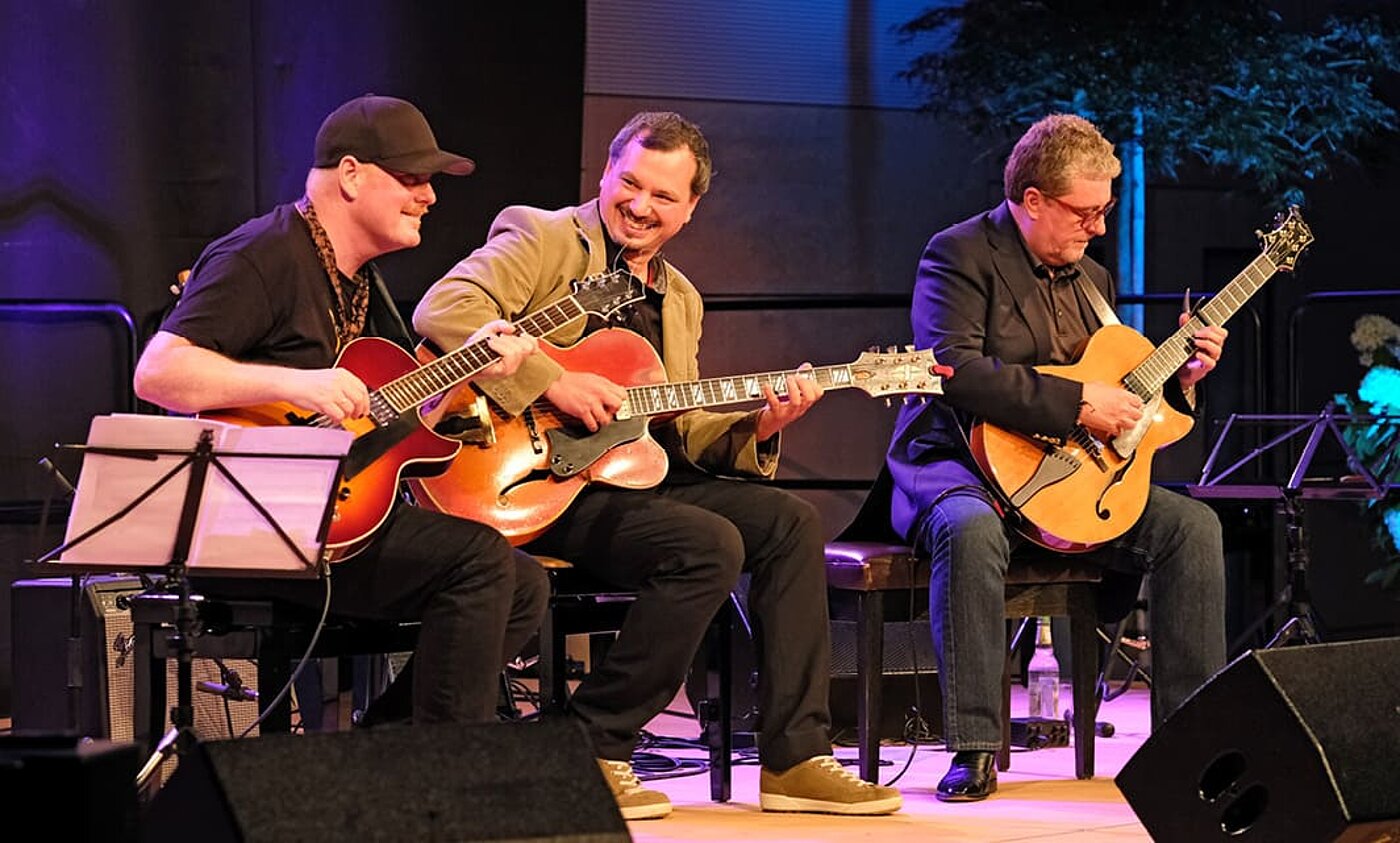 Drei Männer sitzen auf Hockern auf einer Bühne und spielen auf ihren Gitarren. Die Bühne ist bläulich ausgeleuchtet.