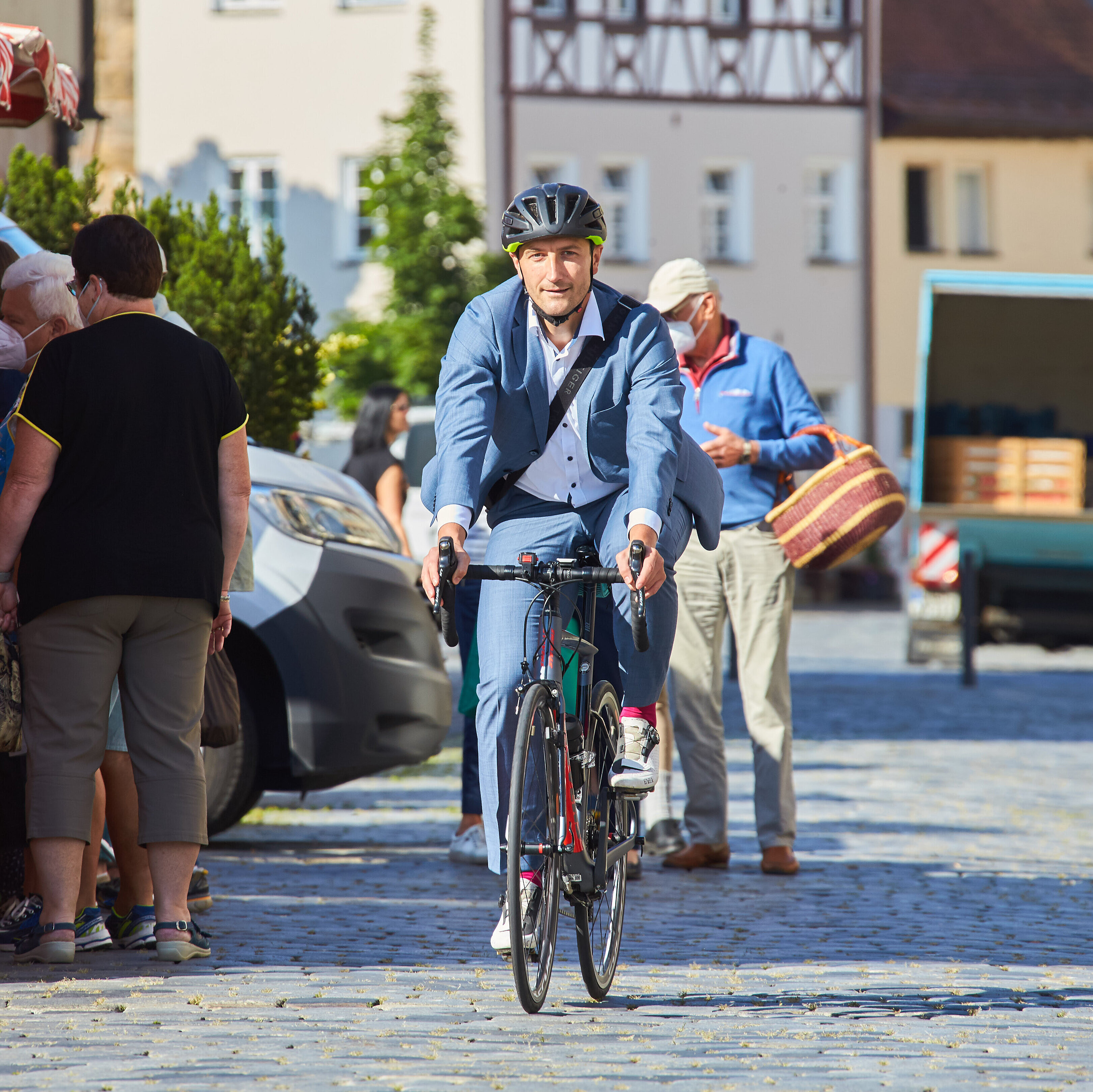 Mann im Anzug fährt mit einem Rennrad über einen Marktplatz.